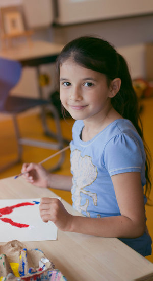 Een meisje met blauw t-shirt en penseel in de hand kijkt lachend naar de camera. Op de tafel voor haar ligt haar schilderij. Foto: Brenda Roos